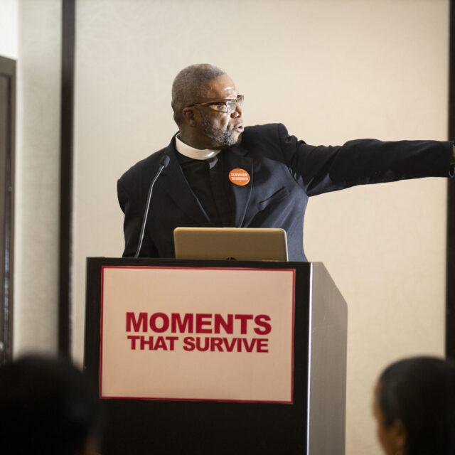 A man at a podium points to something out of frame