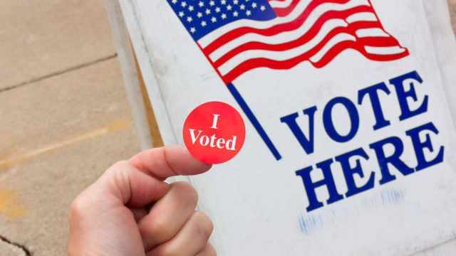 A person holds an I Voted sticker in front of a sign that says Vote Here