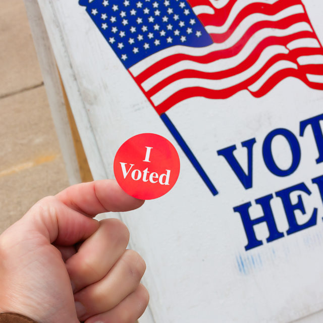 A person holds an I Voted sticker in front of a sign that says Vote Here
