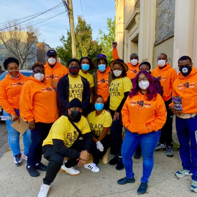A group of 15 people stand in a group together while wearing Paterson Healing Collective sweatshirts and Black Lives Matter shirts. The group is standing outside while wearing masks during the pandemic.