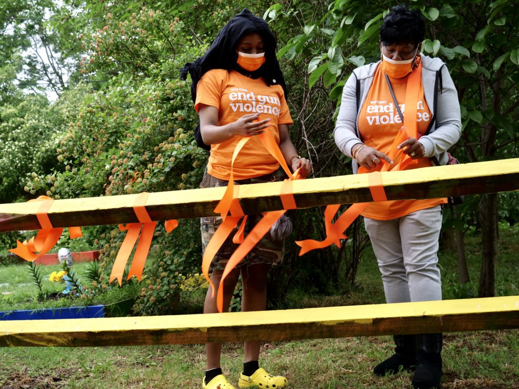 Two volunteers wearing Wear Orange shirts place ribbons on a fence at a Wear Orange event