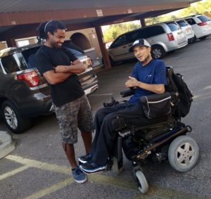 James Hinkley Wade sitting in his wheelchair in a parking lot with a man smiling down at him.