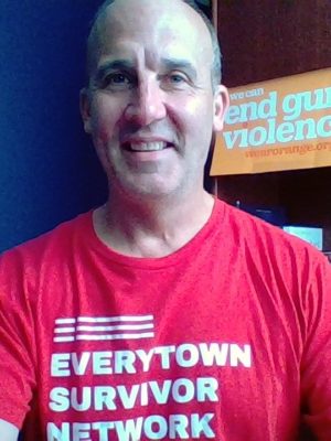 James smiles while wearing his Everytown Survivor Network shirt. Behind him is a sign that reads We Can End Gun Violence.
