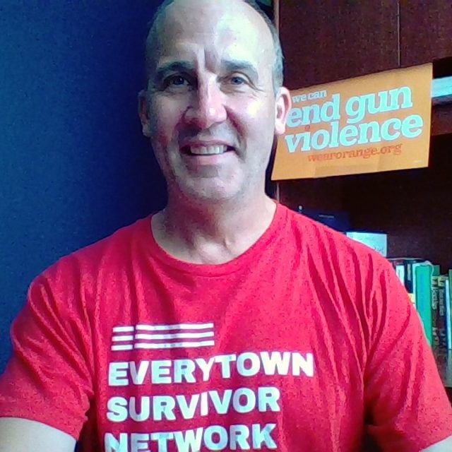 James smiles while wearing his Everytown Survivor Network shirt. Behind him is a sign that reads We Can End Gun Violence.