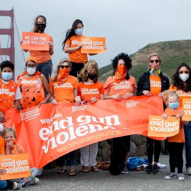 A group of children and adults gathered around a "we can end gun violence" Wear Orange banner, with some individually holding "we can end gun violence" signs
