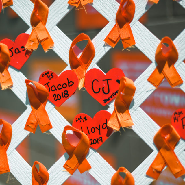 Orange ribbons and paper red hearts with handwritten names and years on them, pasted on a white fence