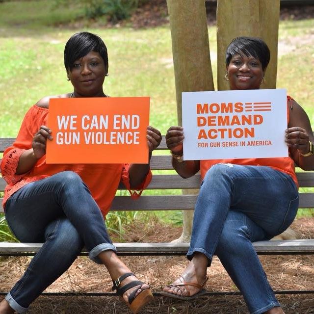 Two women (including Tisa Whack) sit on a park bench holding up signs. One sign is orange and says “We Can End Gun Violence” in white; the other is white and has the Moms Demand Action for Gun Sense in America logo in orange