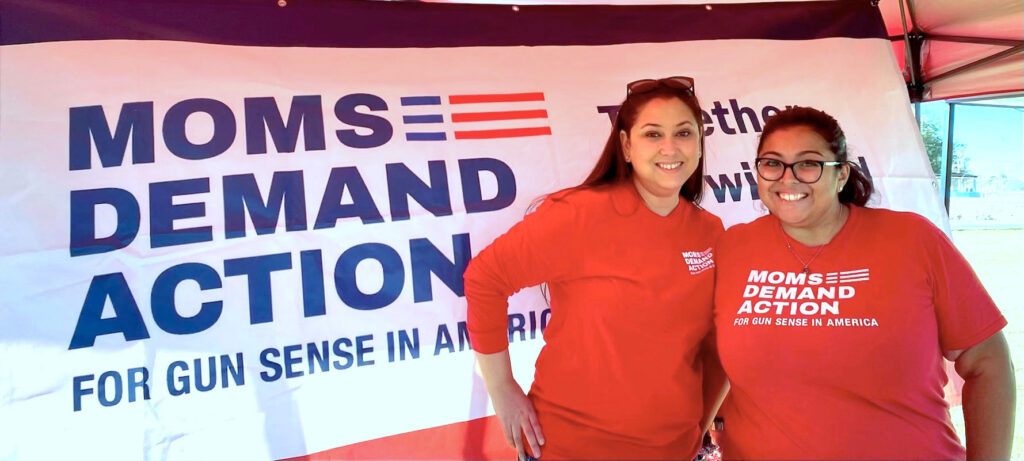 Selina Saenz posing for a photo with another Moms Demand Action volunteer in front of a Moms Demand Action banner