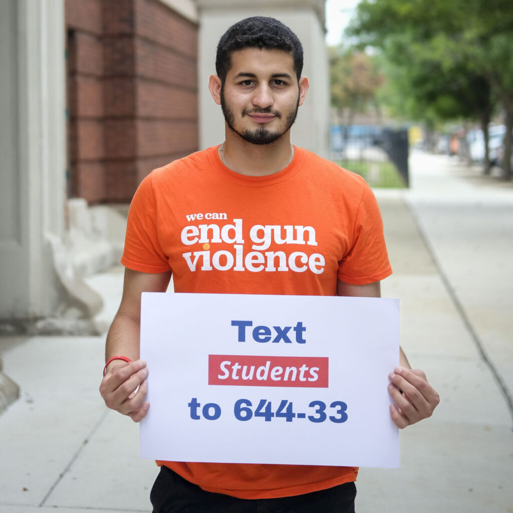 Justin Funez wearing an orange "we can end gun violence" t-shirt and holding a sign that says "Text Students to 644-33"
