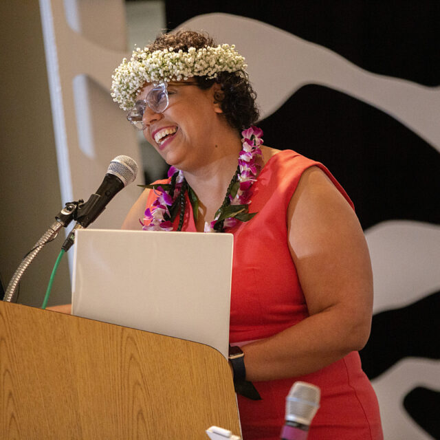 An image of Angelina Mercado, a woman with short, curly dark hair, wearing a white crown of baby's breath and a lei around her neck with magenta and white flowers. She wears a red dress and smiles as she speaks into a microphone at a podium.