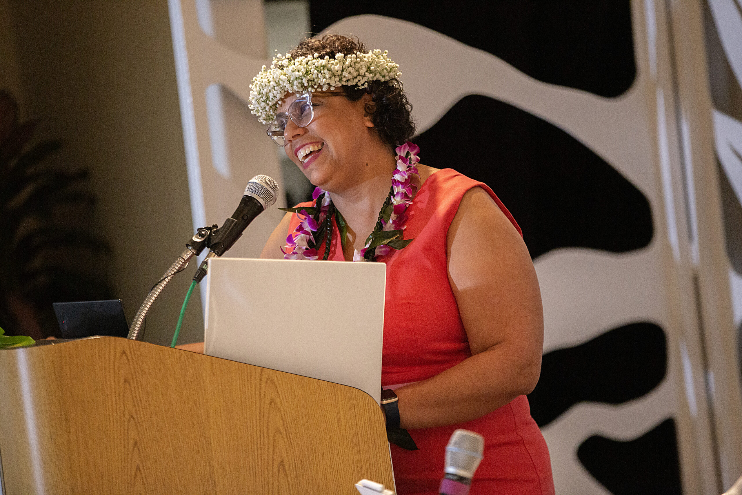 Angelina Mercado, a woman with short, curly dark hair, wears glasses, a crown of baby's breath, and a lei with magenta flowers around her neck. She wears a red dress, a black watch, and smiles from a podium, where she is speaking into a microphone.
