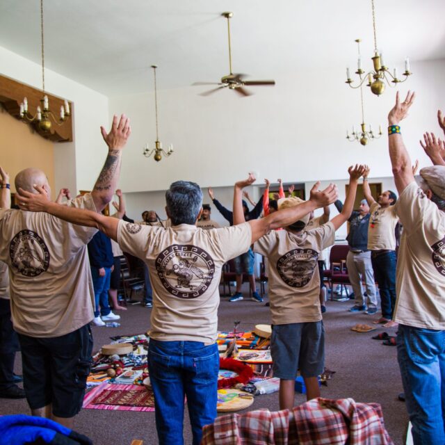 A group of retiro (retreat) participants gather in a circle. They wear denim pants and taupe t-shirts; their arms are stretched up toward the ceiling.
