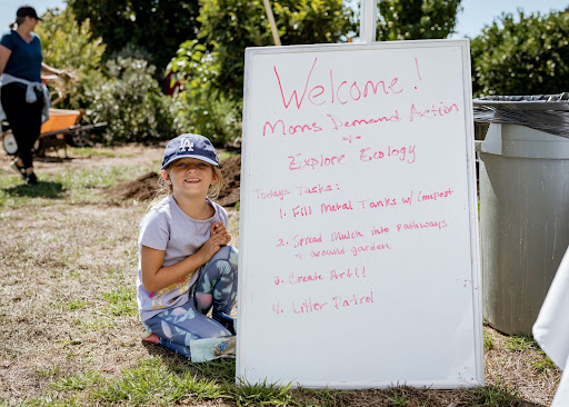 A young girl wearing a blue baseball cap, a light lavender t-shirt, and patterned leggings kneels to the left of a white board that reads "Welcome! Moms Demand Action and Explore Ecology. Today's tasks: 1. Fill metal tanks w/compost; 2. Spread mulch into pathways & around garden; 3. Create Art!!; 4. Litter patrol" in red marker. 