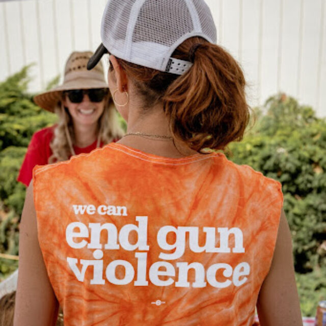 A volunteer is seen from the back. She wears a white mesh baseball cap and has her hair in a short ponytail that brushes the back of her neck. Her shirt is orange tie-dye and reads