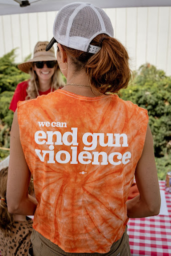 A volunteer is seen from the back. She wears a white mesh baseball cap and has her hair in a short ponytail that brushes the back of her neck. Her shirt is orange tie-dye and reads "we can end gun violence" in white block text.