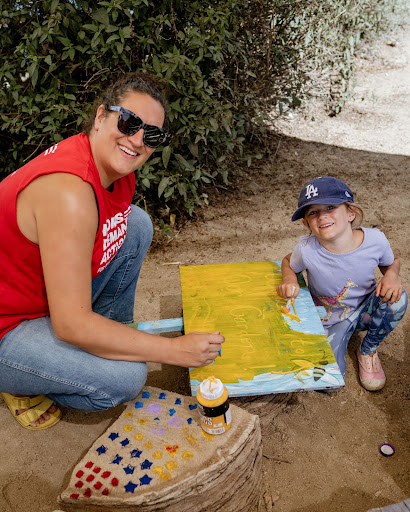 A woman wears blue jeans, black sunglasses, and a sleeveless red t-shirt that reads "Moms Demand Action". She is smiling and kneeling next to a young girl, also smiling and kneeling, who wears a dark blue baseball cap, a purple t-shirt, and leggings. They are painting over an aqua blue sign with yellow paint.