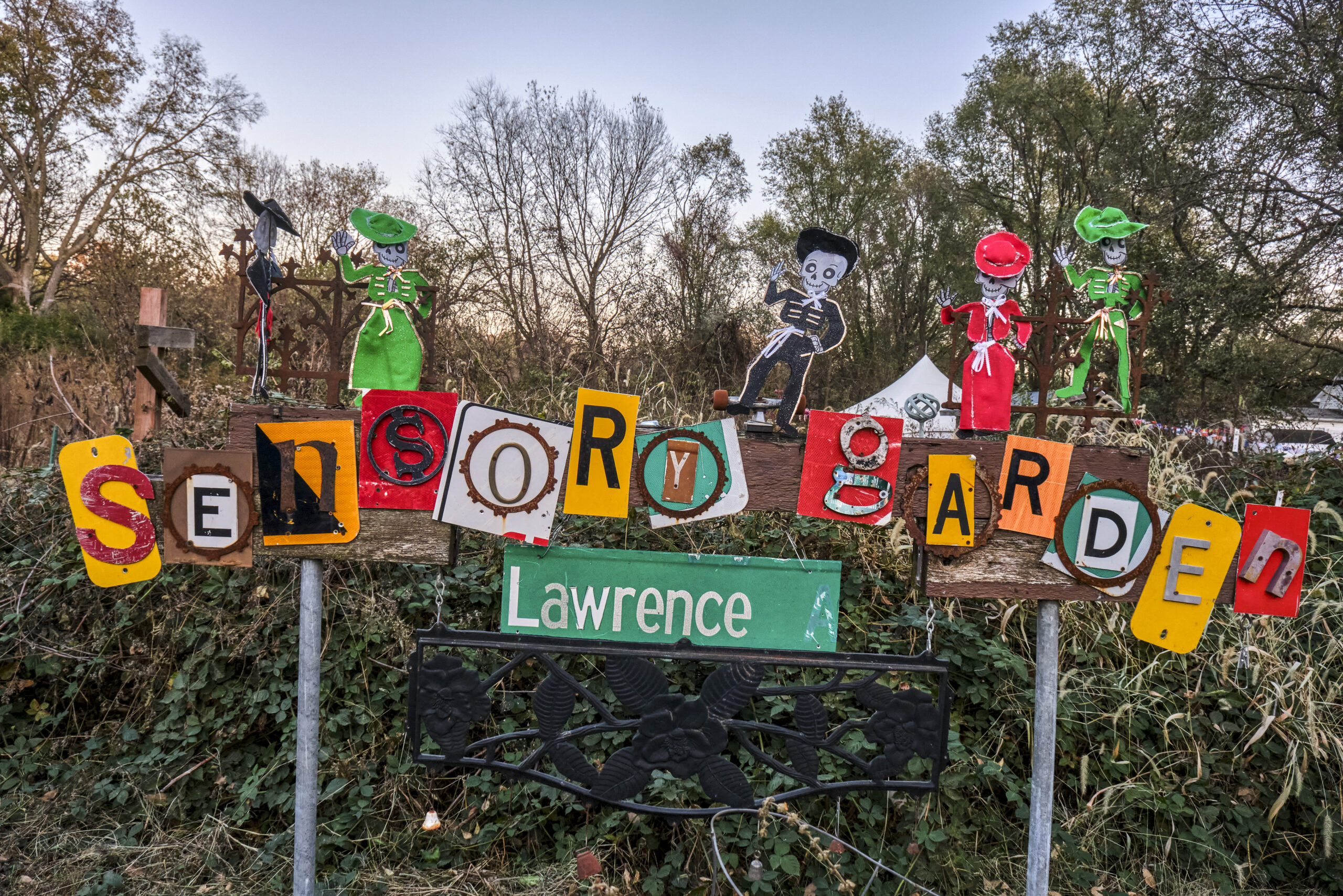 "Sensory Garden" is spelled out in individual block letters on a wooden sign. "Lawrence" is written in white text on a green background, similar to a street sign.