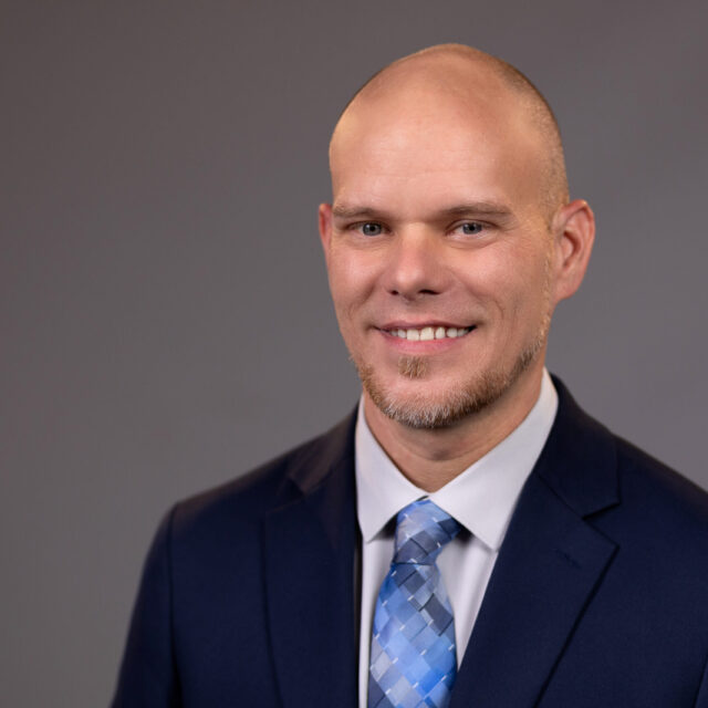 Jeremy Flowers, a white man wearing a navy blue blazer, a white collared shirt, and a gradient blue tie, smiles at the camera. He is pictured in front of a grey professional photo background.