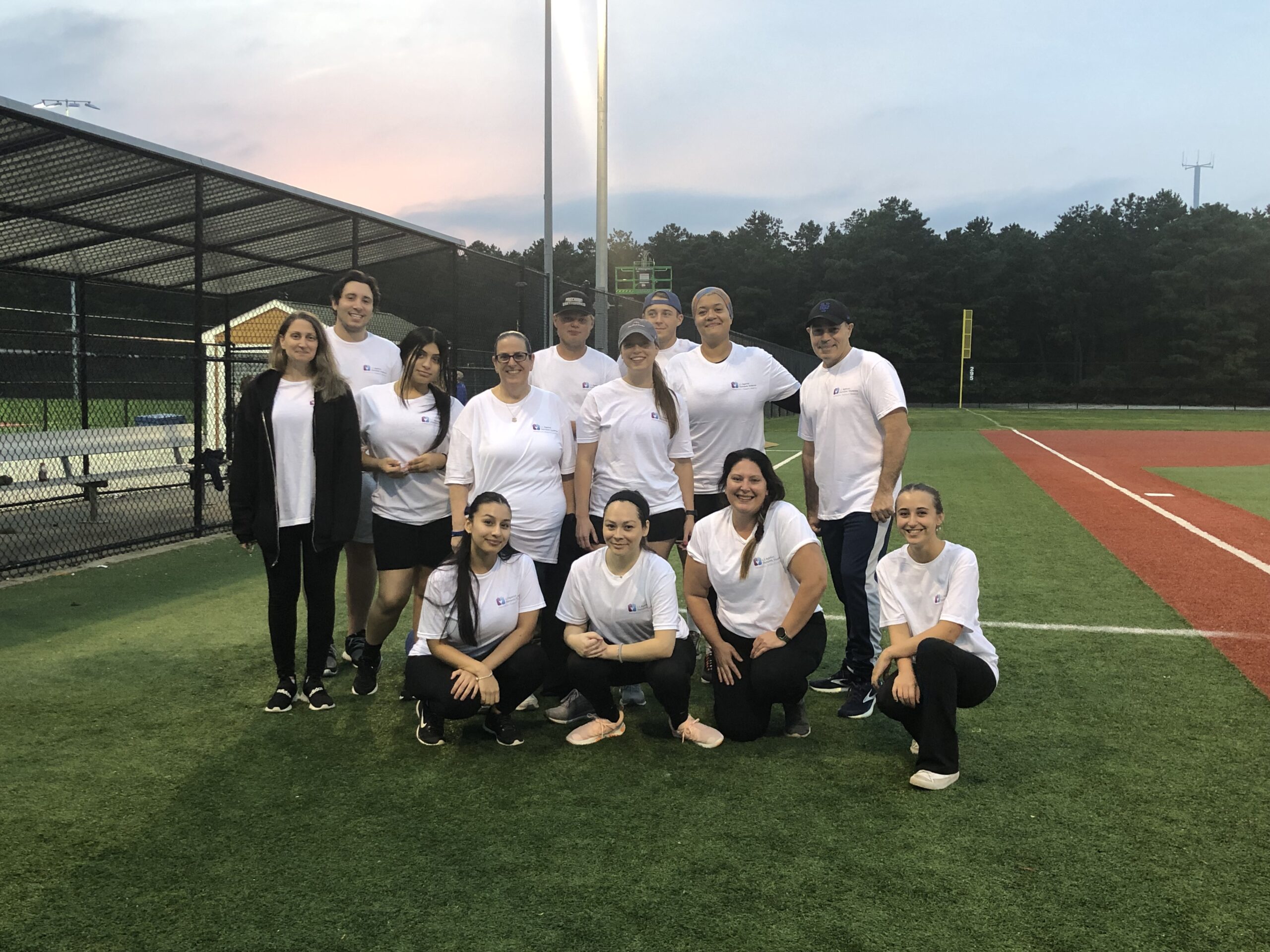  13 members of the LIADV kickball team pose for a photo between a baseball dugout and a baseball diamond. They are all wearing white t-shirts with a small, left-centered LIADV logo