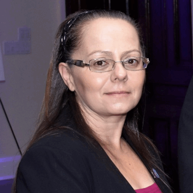 Wendy Linsalata, LIADV Executive Director, poses for a portrait. She wears rectangular glasses with a metal frame, a black blazer, and a maroon blouse. Her straight dark hair is pulled back with a black headband.