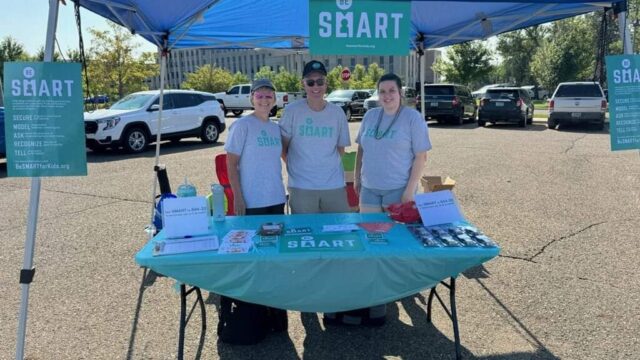 Three Be SMART volunteers table in a parking lot. They wear grey t-shirts with the teal Be SMART logo.