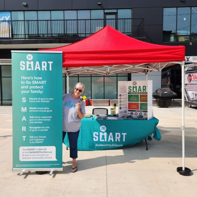 A Be SMART volunteer wears black pants and a grey t-shirt with the teal Be SMART logo. She stands next to a teal Be SMART standard. Behind her is a table covered with a teal tablecloth with the Be SMART logo and a red canopy to give shade while tabling.