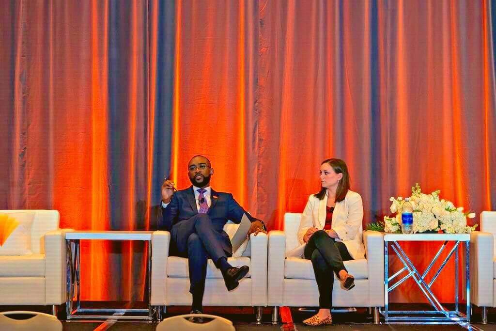 Michael-Sean Spence and Christa Green sit in white armchairs on a stage with an orange-red curtain behind them. Michael-Sean is speaking, holding papers in his left hand and gesturing with his right while talking to the audience. Christa is looking at Michael-Sean; her hands rest in her lap and her right leg is crossed over her left.