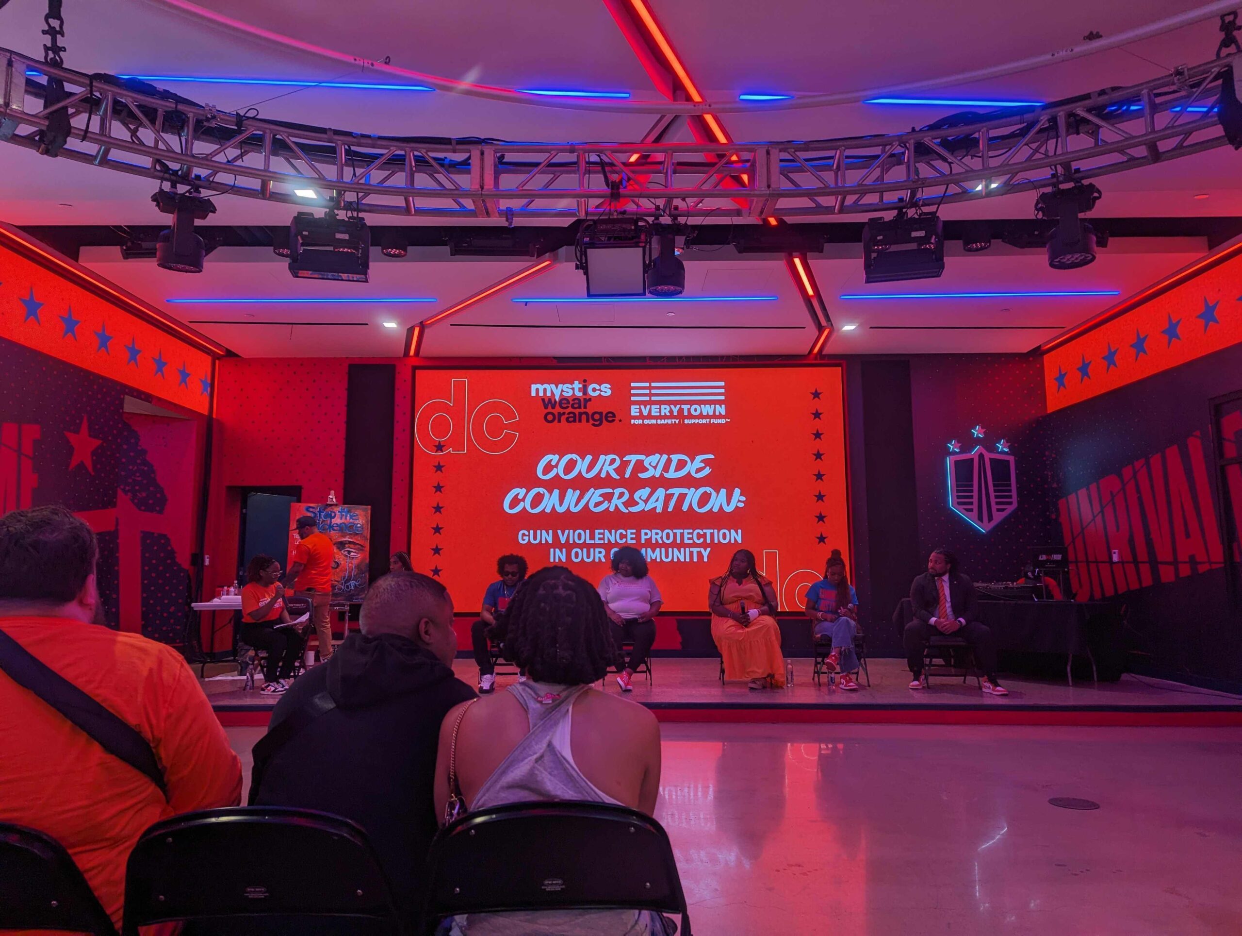 Six panelists participate in a discussion facilitated by Nathalie Arzu. Behind the panelists, an orange-hued slide has two logos at the top: Mystics Wear Orange and Everytown for Gun Safety Support Fund. The slide reads "Courtside Conversation: Gun Violence Prevention in Our Communities."