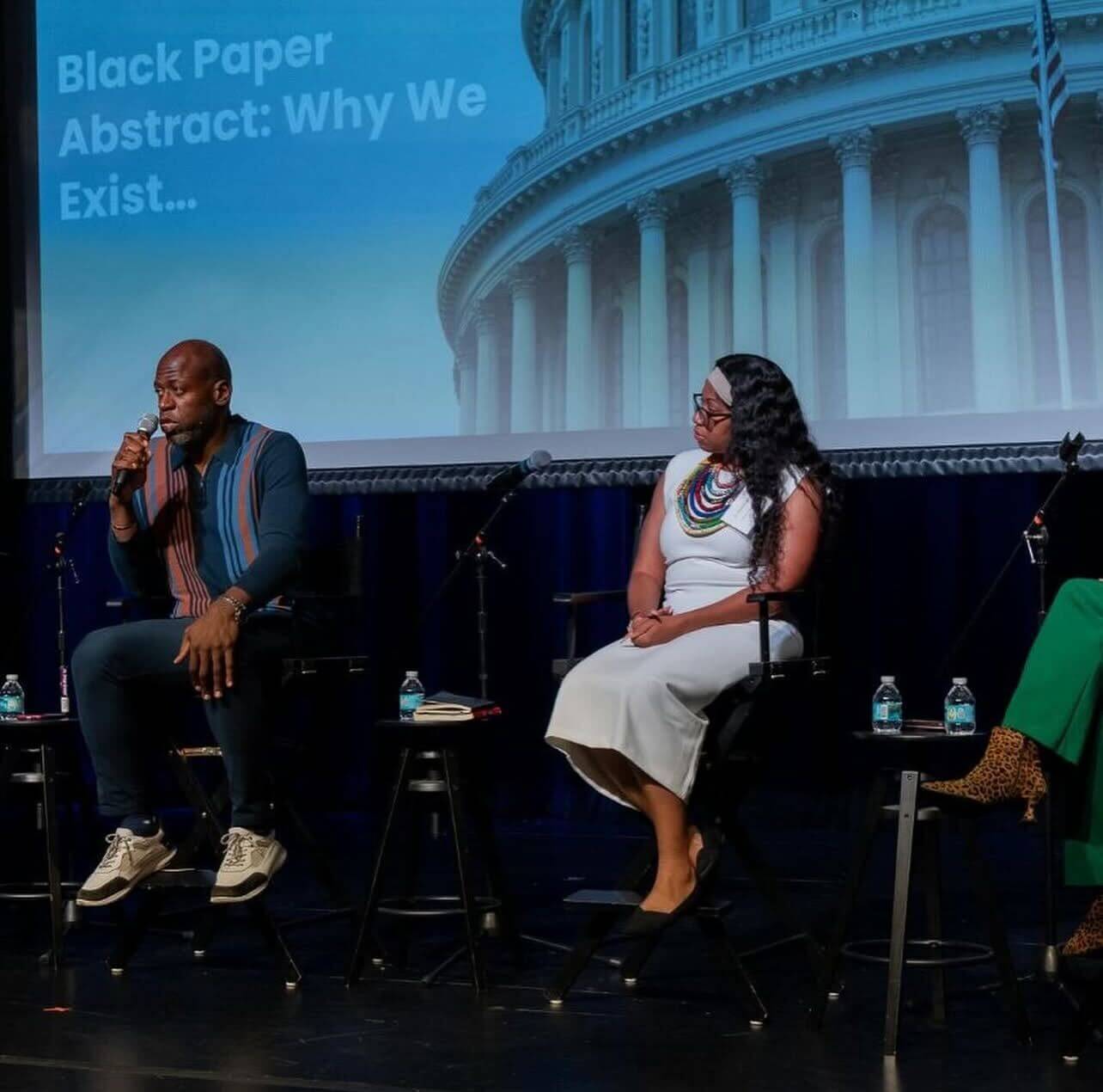 Two panelists speak during a discussion at the State of Emergency convening in Miami, Florida. One is a man wearing black, white, and tan sneakers, navy pants, and a navy shirt with vertical bright blue, cobalt blue, and coral stripes. The other is a woman wearing a white dress, black shoes, and a rainbow-colored multi-strand necklace.