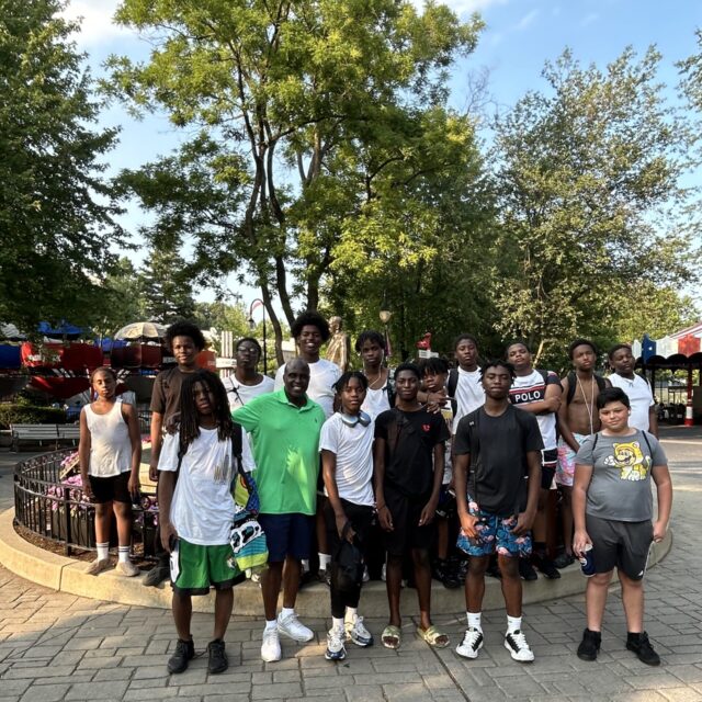 A group of 15 mentees post with a ManUpPHL mentor. They appear to be at an amusement park. The mentees are boys between the ages of 13 and 17.