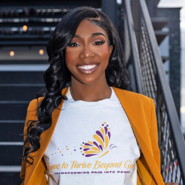 Aaliyah Strong, the founder and executive director of Tyme to Thrive Beyond Grief, poses for a portrait photo. Her black hair is parted on the side and she is wearing subtle glamorous makeup. She is facing the camera directly and is pulling her orange blazer slightly away from her white t-shirt so the full 