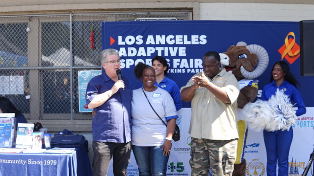 Jamie Christian, the executive director of A New Beginning 4 You Foundation, is pictured in a candid photo at the 2025 Los Angeles Adaptive Sports Fair. She is standing between two event organizers, who are speaking into microphones to an off-camera group of people.