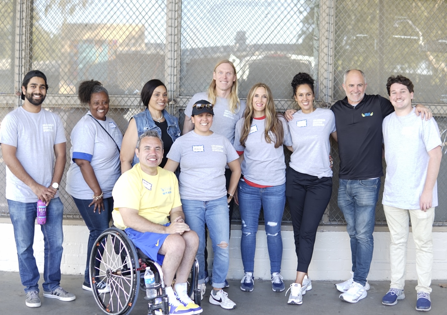 Organizers of the Los Angeles Adaptive Sports Fair pose for a photo and smile. There are 10 people in total, 9 of whom are standing in a row and one of whom is sitting in a wheelchair.