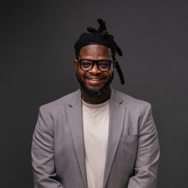 A portrait photo of DuJuan Kennedy, who is posing in front of a professional charcoal-gray background. He wears glasses with a thick black frame, a cream-colored t-shirt, and a thin plaid-stripe blazer.