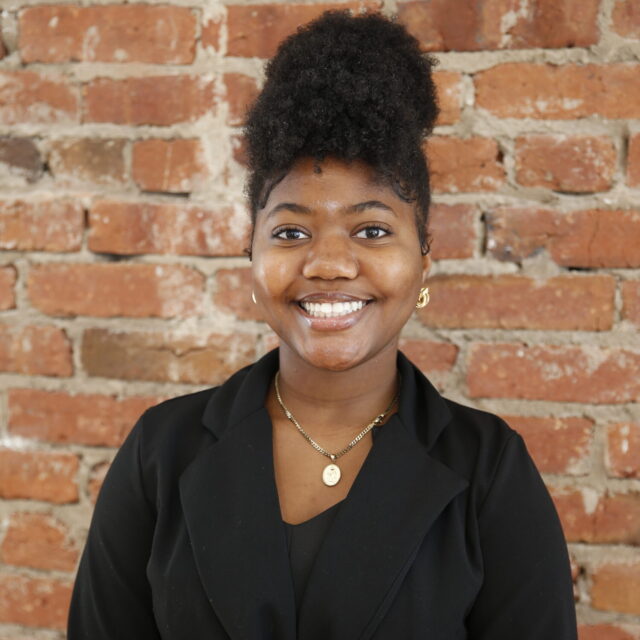 Iyanah Forbes, an HBCU student and the chartering president of Students Demand Action, poses for a professional photo. She stands in front of a brick wall and wears a black blazer, a gold necklace with a pendant, and small dangly gold earrings.