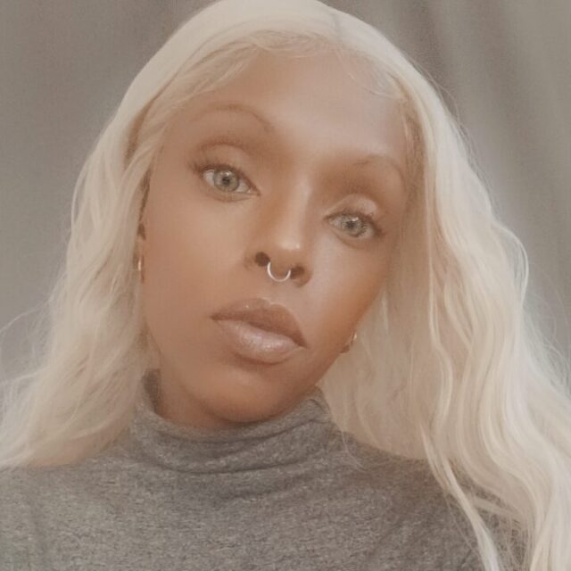 Marquita Wisley, a Black woman, takes a selfie. She is wearing a grey turtleneck sweater and a large aqua blue ring with a silver setting.
