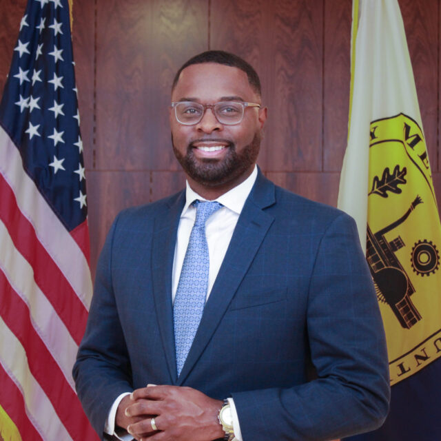 City of Memphis Paul A. Young poses for a professional photo. He is wearing a navy suit jacket, a white collared shirt, and a blue patterned tie. He wears glasses with a light frame and a metal watch. The background is a wood-paneled wall, and he stands between the American flag and the flag of Memphis, Tennessee.