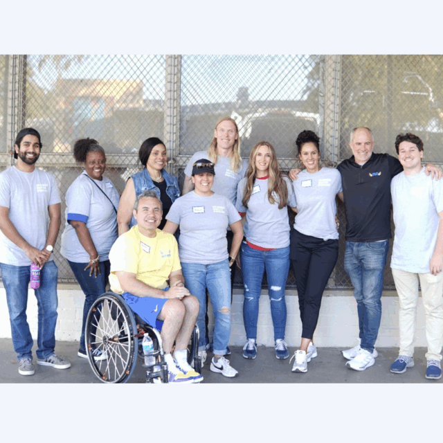Organizers of the Los Angeles Adaptive Sports Fair pose for a photo and smile. There are 10 people in total, 9 of whom are standing in a row and one of whom is sitting in a wheelchair.