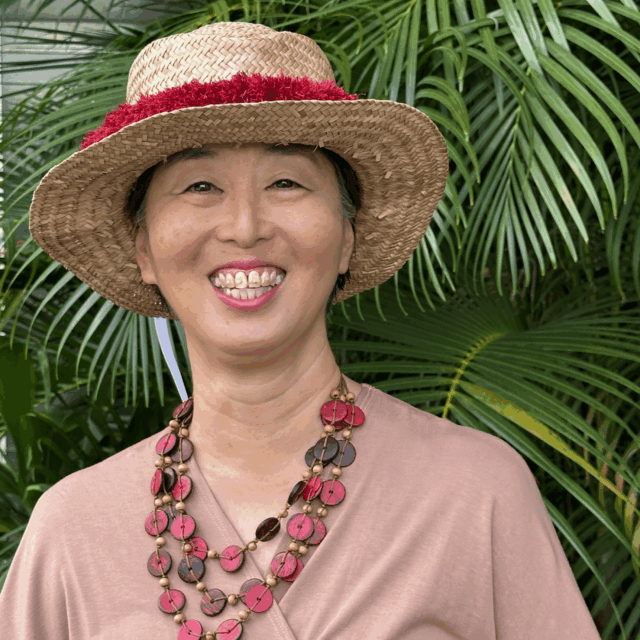 A photo of Elna Nagasako from the waist up, who is pictured in front of some ferns while wearing a dusky rose-colored pink wrap top, a chunky multi-strand necklace, and a straw hat.