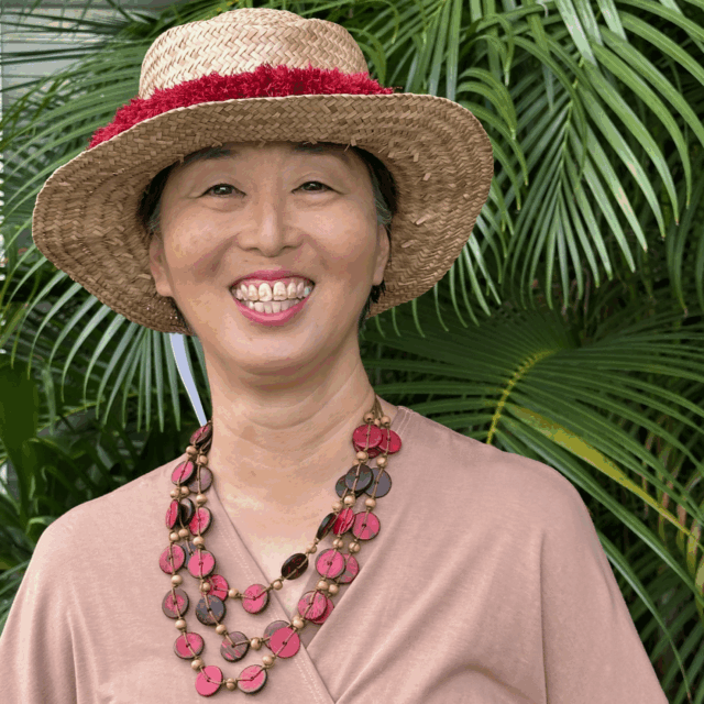 A photo of Elna Nagasako from the waist up, who is pictured in front of some ferns while wearing a dusky rose-colored pink wrap top, a chunky multi-strand necklace, and a straw hat.