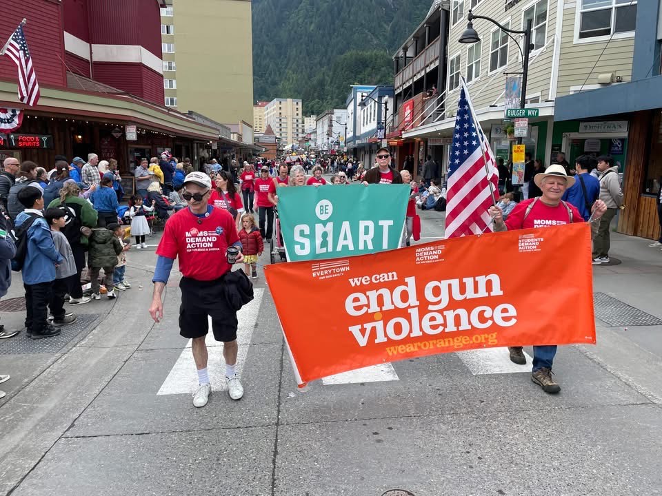 Alaska Moms Demand Action volunteers walk in a town parade. They are wearing red Moms Demand Action t-shirts and carry banners that say "Be SMART" and "we can end gun violence."