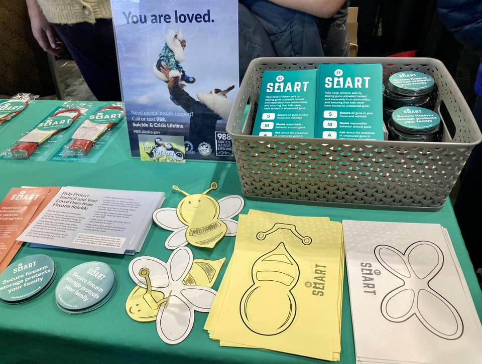Be SMART materials displayed on a table at a community event, including coloring pages, a poster that reads "you are loved" with information for the 988 Suicide & Crisis Lifeline, and Be SMART stickers.