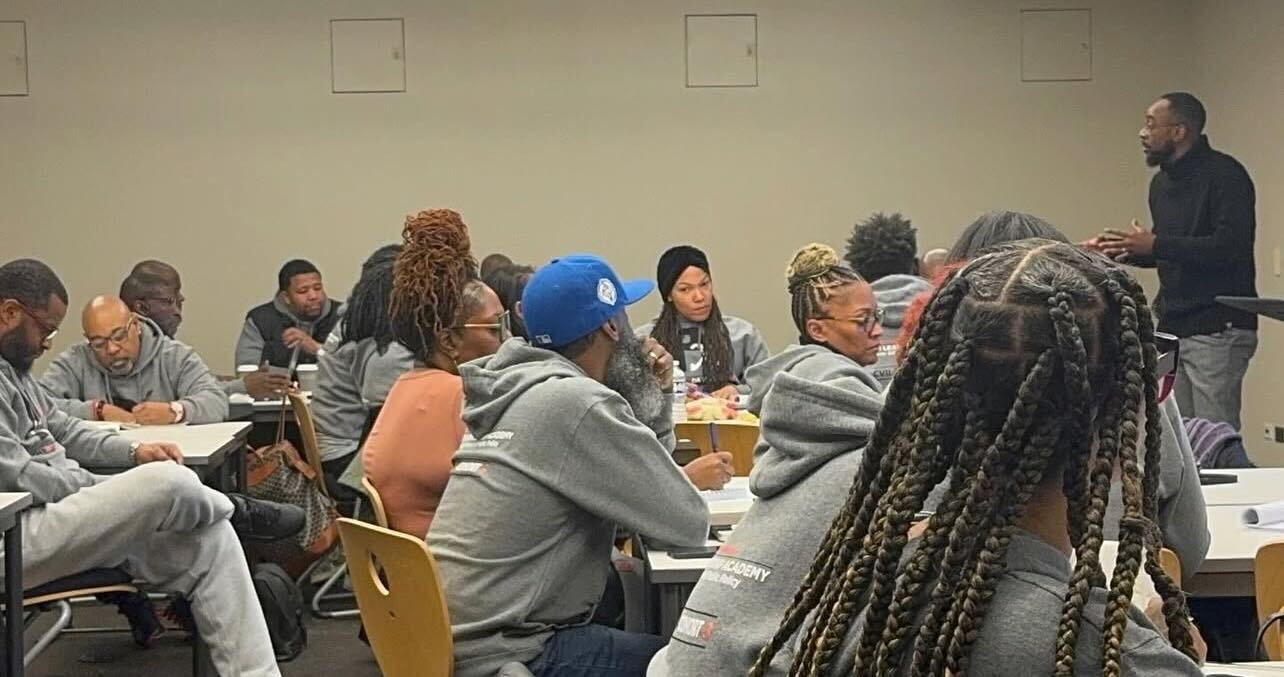 A candid photo of Michael-Sean Spence, a Black man wearing light dress pants and a dark long-sleeve shirt, teaching a room full of community violence intervention practitioners during a training at the University of Chicago's Community Violence Intervention Leadership Academy.